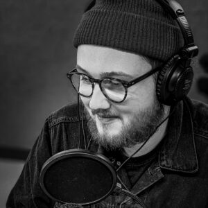 A man wearing a beanie hat, headones and glasses talking in to a microphone with a pop shield